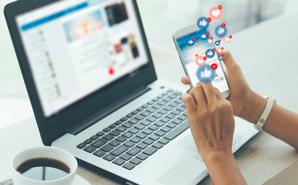 Business person using workplace social media on a phone in front of a work laptop.